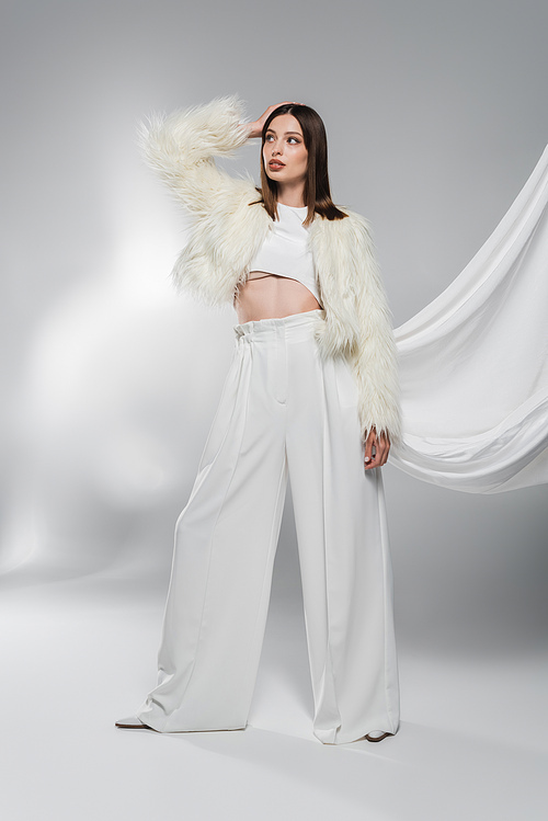 Trendy woman in faux fur jacket and white pants posing on abstract grey background