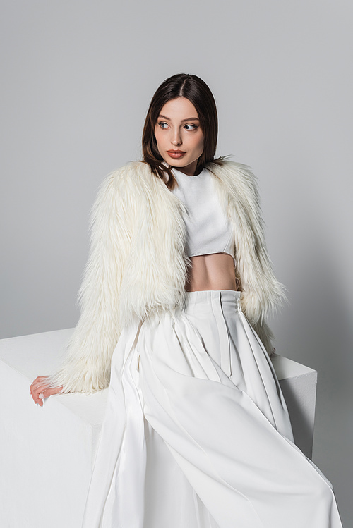 trendy young woman in faux fur jacket and total white outfit leaning on cube on grey