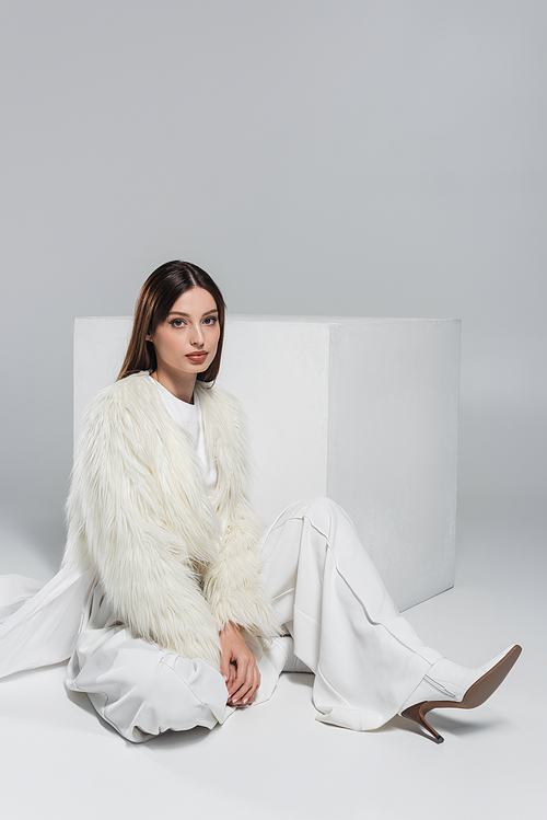 full length of trendy woman in faux fur jacket and total white outfit sitting near cube on grey