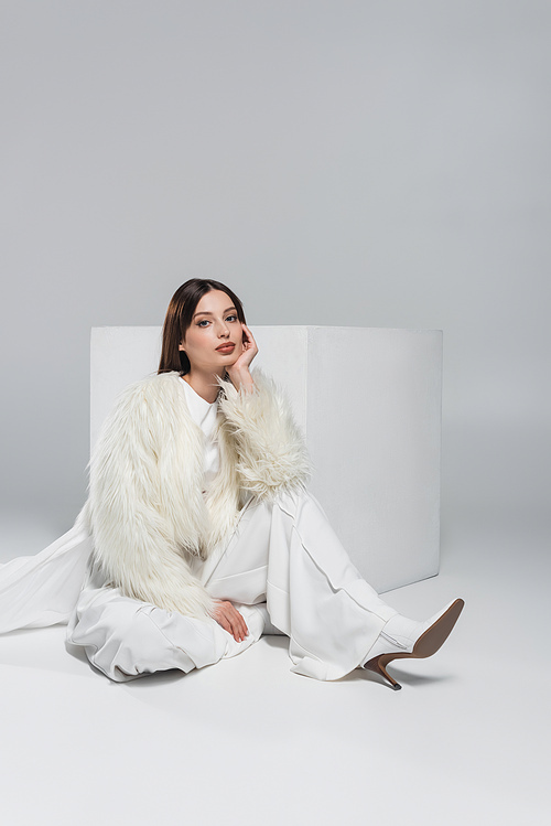 full length of young woman in faux fur jacket and total white outfit sitting near cube on grey