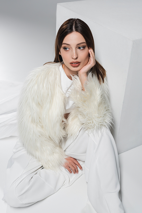 high angle view of young woman in faux fur jacket and total white outfit sitting near cube on grey