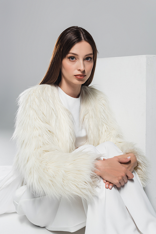 stylish young woman in white faux fur jacket looking at camera near cube on grey