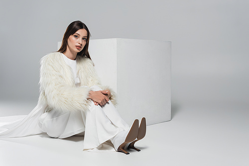 full length of stylish young woman in white faux fur jacket looking at camera near cube on grey background