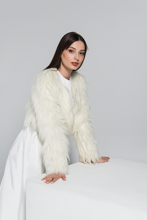 young woman in faux fur jacket looking at camera and leaning on white cube isolated on grey