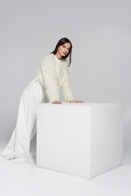 full length of stylish young woman in faux fur jacket looking at camera while leaning on white cube on grey