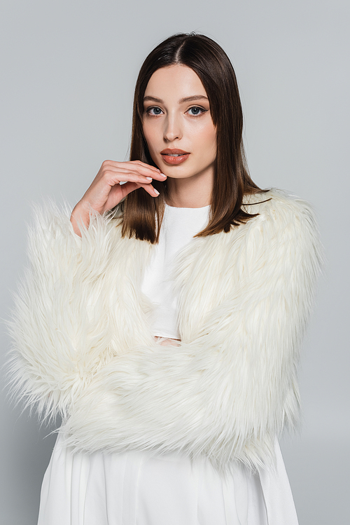 portrait of pretty young woman in stylish white faux fur jacket looking at camera isolated on grey