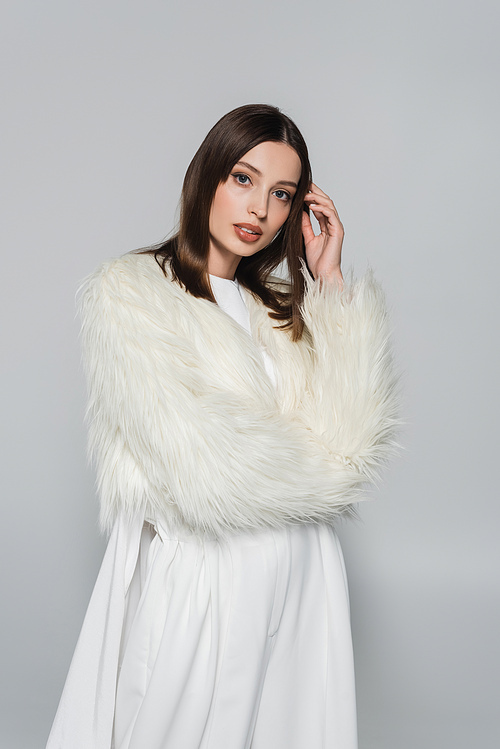 portrait of stylish young woman in white faux fur jacket looking at camera isolated on grey