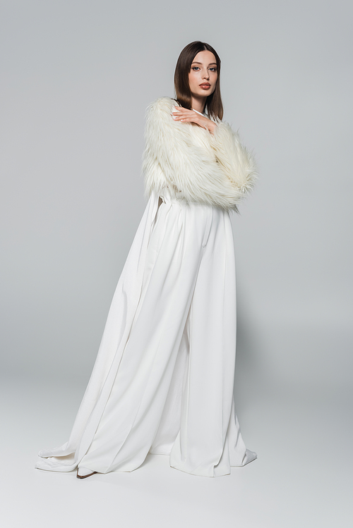 full length of trendy woman in totally white outfit and faux fur jacket posing on grey