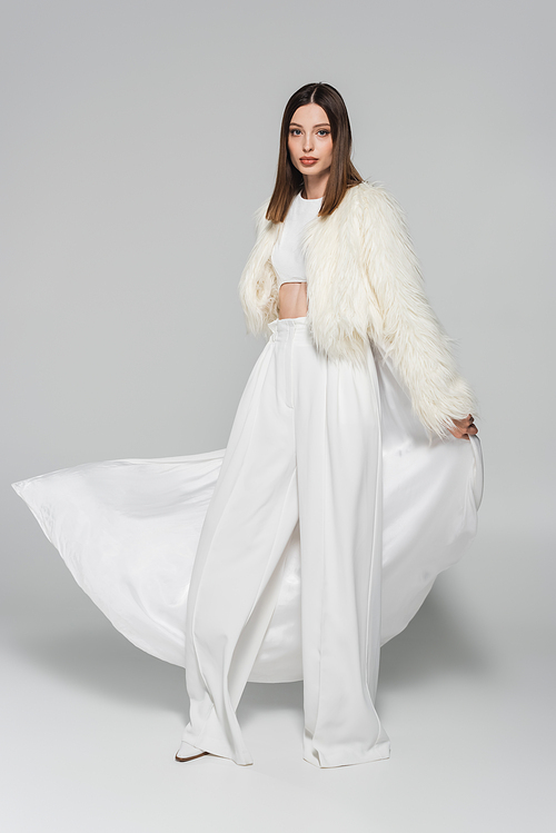 full length of trendy woman in totally white outfit and faux fur jacket standing on grey