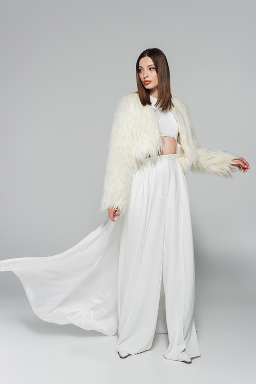 full length of young woman in totally white outfit and trendy faux fur jacket posing on grey