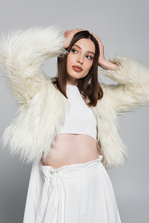 young woman in totally white outfit and trendy faux fur jacket posing with hands near head isolated on grey