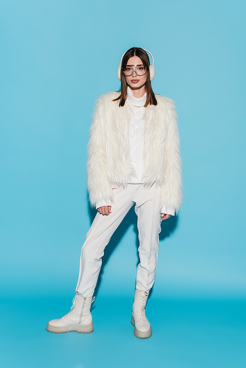 full length of stylish woman in earmuffs and eyeglasses posing in faux fur jacket on blue