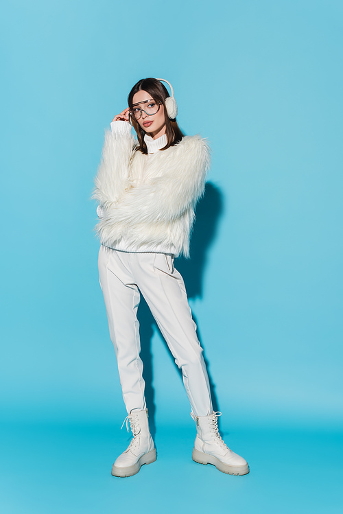 full length of stylish young woman in earmuffs adjusting trendy eyeglasses while posing in faux fur jacket on blue