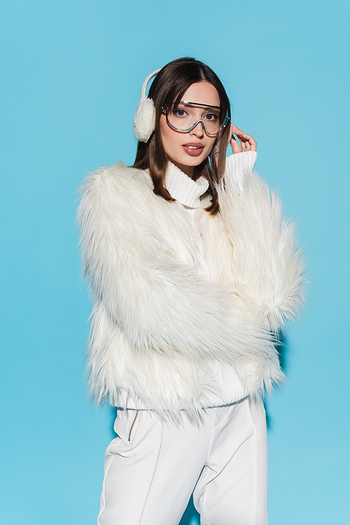 stylish young woman in winter earmuffs and eyeglasses posing in white faux fur jacket on blue