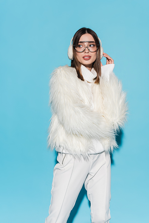 stylish young woman in winter earmuffs and trendy eyeglasses posing in white faux fur jacket on blue