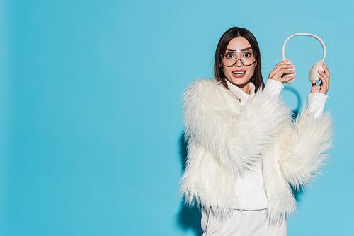 amazed young woman in eyeglasses and stylish faux fur jacket holding winter earmuffs on blue