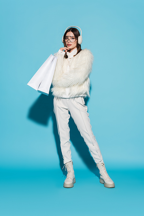 full length of stylish woman in faux fur jacket and eyeglasses holding shopping bags on blue