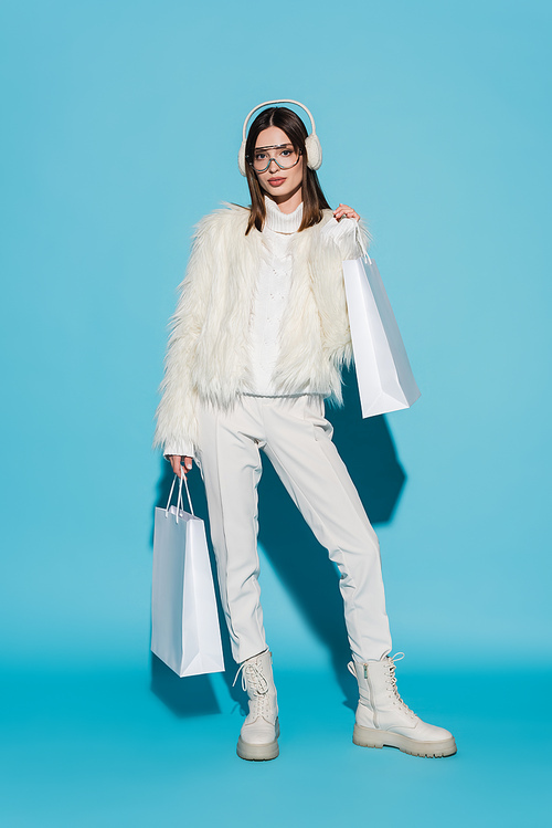 full length of young woman in earmuffs and stylish faux fur jacket holding shopping bags on blue