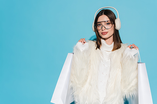 young woman in earmuffs and stylish faux fur jacket holding shopping bags on blue background