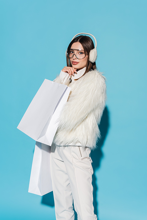 young woman in winter earmuffs and trendy faux fur jacket holding shopping bags on blue