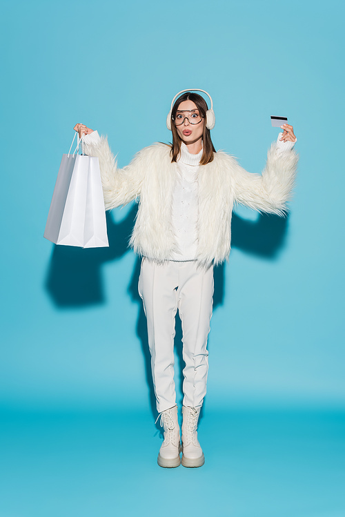 full length of surprised woman in eyeglasses and stylish faux fur jacket holding credit card and shopping bags on blue