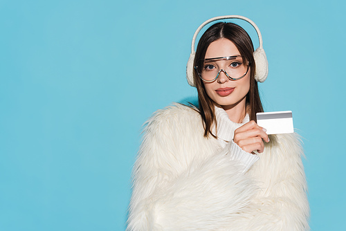 pleased young woman in eyeglasses and stylish faux fur jacket holding credit card isolated on blue