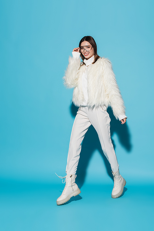 full length of cheerful woman in faux fur jacket and stylish eyeglasses walking on blue