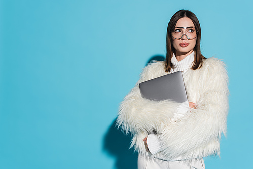 stylish woman in trendy eyeglasses and stylish faux fur jacket holding laptop on blue background