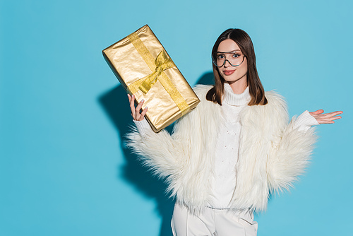 stylish woman in faux fur jacket and eyeglasses holding christmas present on blue background