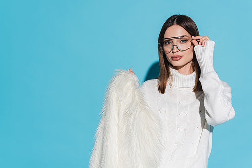 stylish young woman adjusting trendy eyeglasses and holding white faux fur jacket on blue