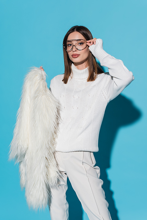 pretty and young woman adjusting trendy eyeglasses while holding white faux fur jacket on blue