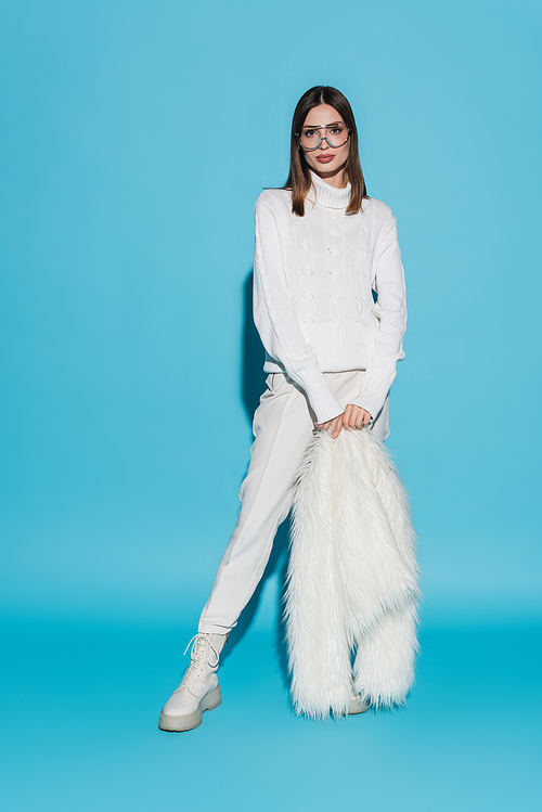 full length of stylish young woman in trendy eyeglasses and turtleneck holding white faux fur jacket on blue