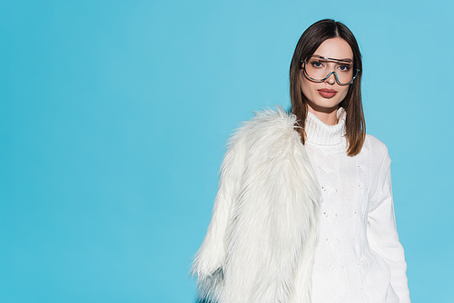 stylish young woman in trendy eyeglasses and turtleneck holding white faux fur jacket on blue