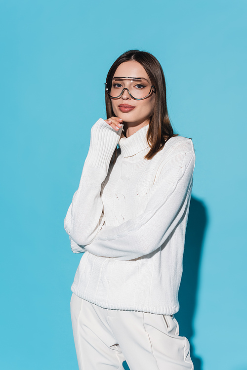 stylish young woman in eyeglasses and white turtleneck looking at camera on blue