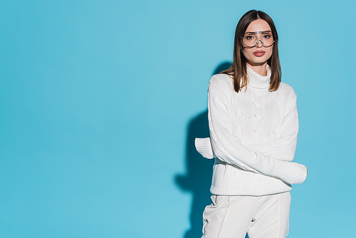 pretty young woman in trendy eyeglasses and white turtleneck posing with crossed arms on blue background