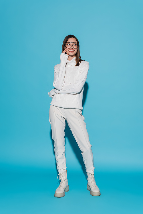 full length of joyful young woman in eyeglasses and totally white outfit posing on blue