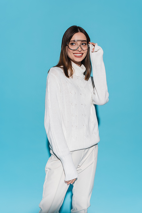joyful young woman in trendy eyeglasses and totally white outfit posing on blue