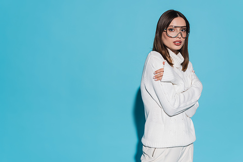 pretty young woman in trendy eyeglasses and totally white outfit posing with crossed arms on blue