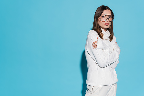 pretty young woman in trendy eyeglasses and white outfit posing with crossed arms on blue