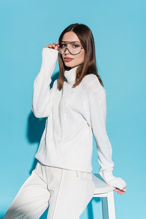 young and stylish woman in white outfit adjusting eyeglasses while leaning on high chair on blue