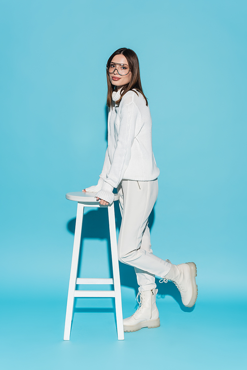 full length of trendy model in white outfit and eyeglasses leaning on high chair on blue
