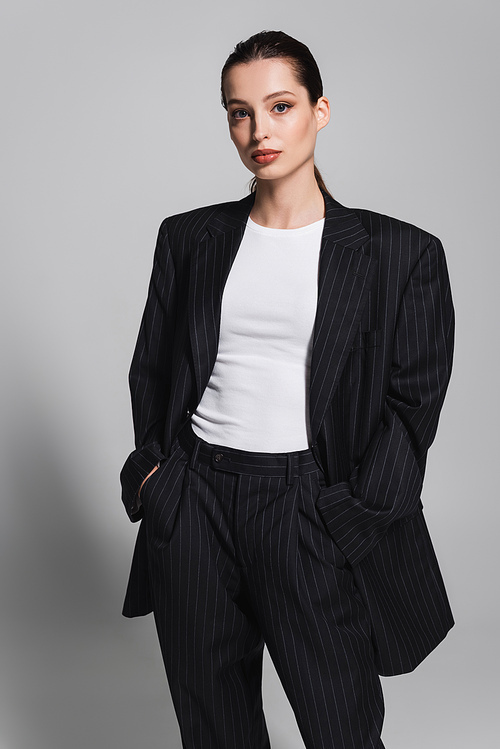 Trendy woman in black suit posing with hands in pockets on grey background