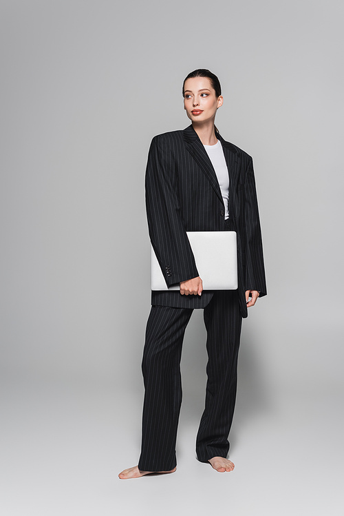 Stylish barefoot woman in suit holding laptop and looking away on grey background