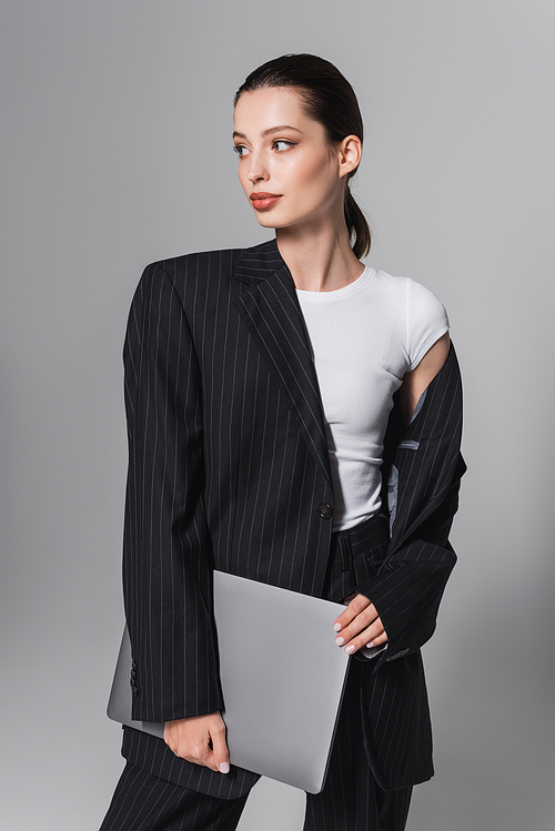 Pretty brunette woman in striped suit holding laptop isolated on grey