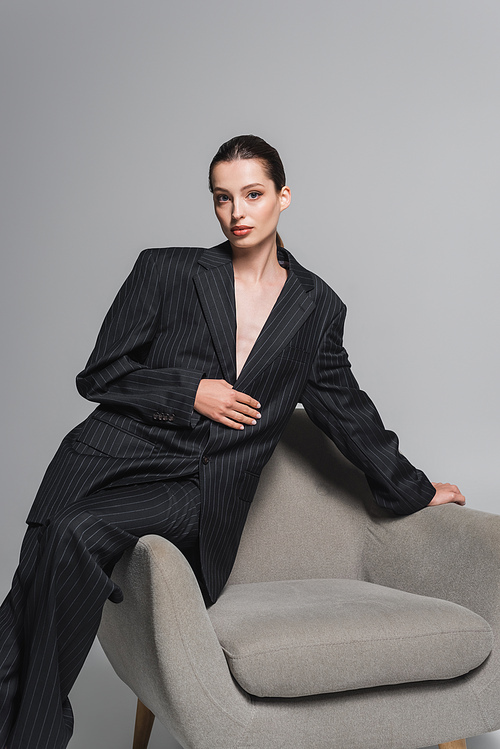 Fashionable woman in suit posing near armchair on grey background