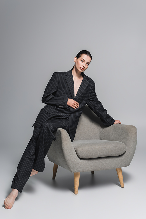 Full length of trendy young woman in suit posing near armchair on grey background