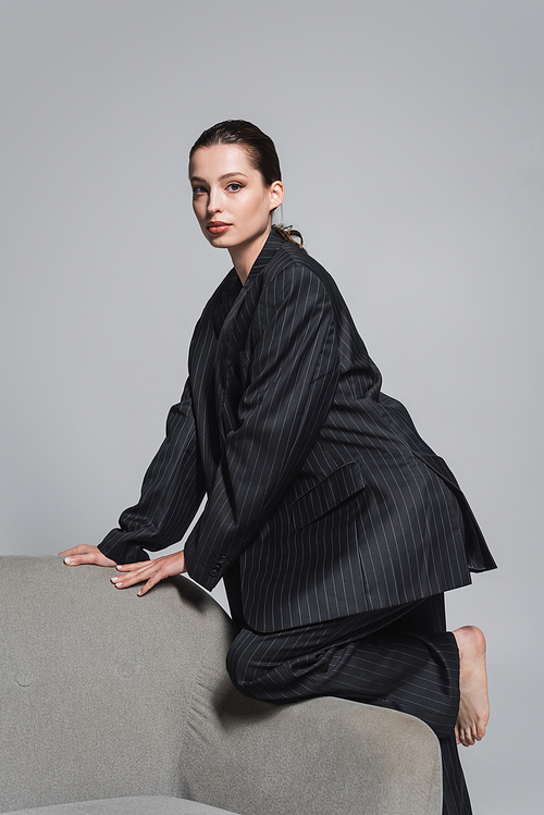 Brunette woman in striped suit posing near armchair isolated on grey