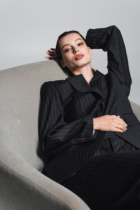 Stylish woman in black suit posing on armchair isolated on grey