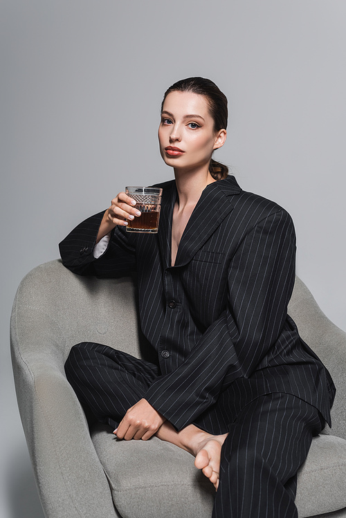 Stylish model in black suit holding glass of whiskey while sitting on armchair on grey background