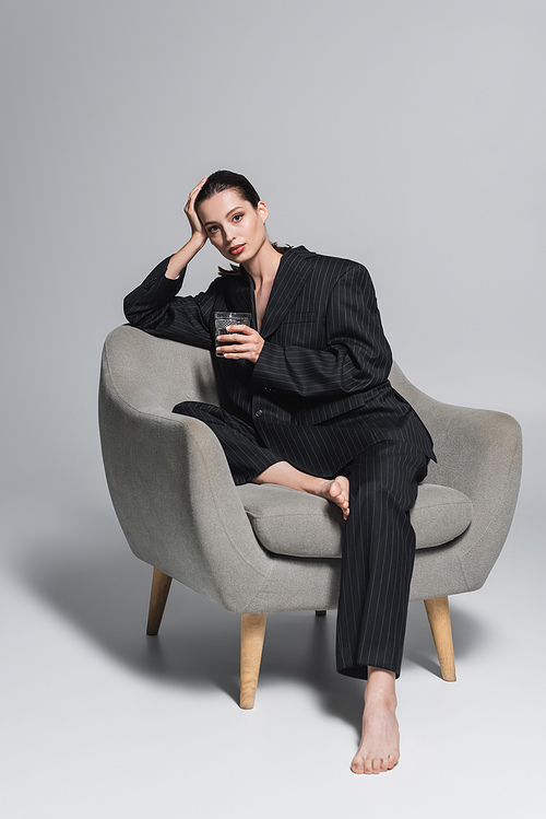 Barefoot woman in black suit holding glass of whiskey while sitting on armchair on grey background